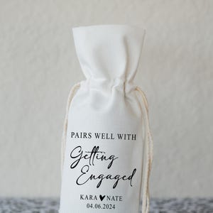 Op de afbeelding: Een witte stoffen tas met een trekkoord. De tas heeft zwarte tekst die "Pairs well with Getting Engaged Kara &hearts; Nate 04.06.2024" luidt.