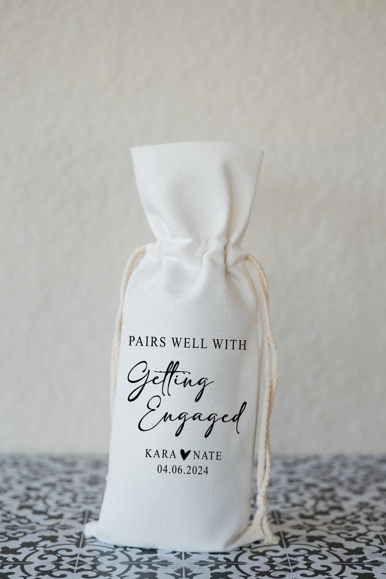 Op de afbeelding: Een witte stoffen tas met een trekkoord. De tas heeft zwarte tekst die "Pairs well with Getting Engaged Kara &hearts; Nate 04.06.2024" luidt.