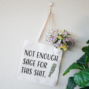 May include: White tote bag with black text that reads "Not Enough Sage For This Shit." A small illustration of a sage bundle is next to the text. The bag is hanging from a hook on a white wall. A bouquet of flowers is hanging from the bag.