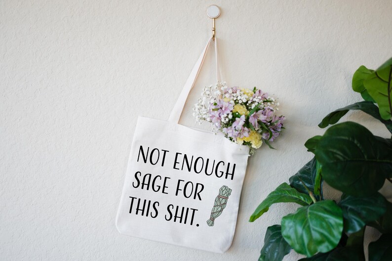 May include: White tote bag with black text that reads "Not Enough Sage For This Shit." A small illustration of a sage bundle is next to the text. The bag is hanging from a hook on a white wall. A bouquet of flowers is hanging from the bag.