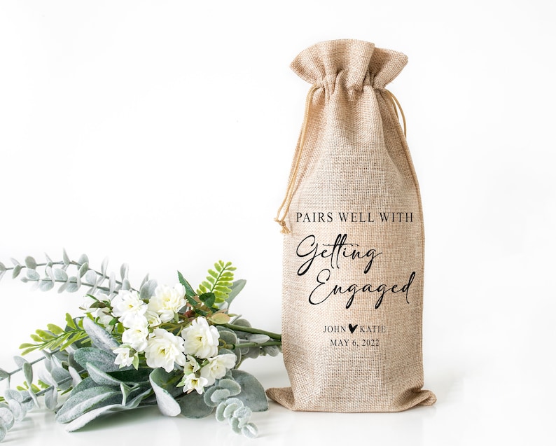 Op de afbeelding: Een jute wijnfleszak met de tekst "Pairs well with Getting Engaged John &hearts; Katie May 6, 2022".