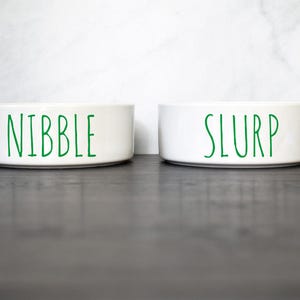 May include: Two white ceramic pet bowls with green lettering. The bowl on the left says "Nibble" and the bowl on the right says "Slurp".