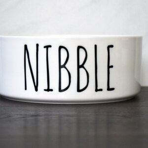May include: White ceramic pet bowl with the word "NIBBLE" printed in black.