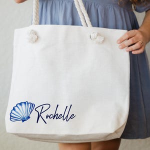 May include: White canvas tote bag with rope handles. The bag has a blue seashell and the name "Rochelle" printed on it.