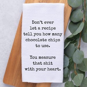 May include: White waffle-weave kitchen towel with black text: "Don't ever let a recipe tell you how many chocolate chips to use. You measure that shit with your heart." The towel rests on a wooden cutting board.