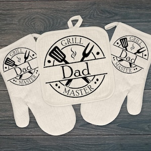 May include: Off-white grilling set featuring a pot holder and two oven mitts. Each item has a black circular design with the words "GRILL Dad MASTER" and images of a spatula, fork, and flame.