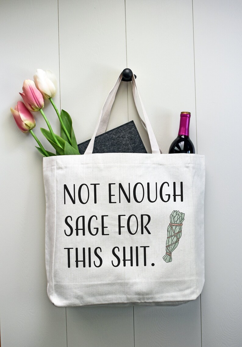 May include: White canvas tote bag with black text that reads "Not Enough Sage For This Shit." A small illustration of a bundle of sage is to the right of the text. A bottle of wine and a bouquet of pink tulips are in the bag.