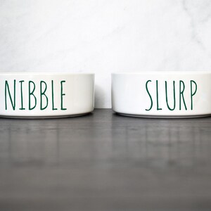 May include: Two white ceramic pet bowls with green lettering. The bowl on the left says "Nibble" and the bowl on the right says "Slurp".