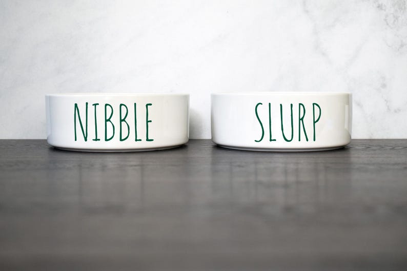 May include: Two white ceramic pet bowls with green lettering. The bowl on the left says "Nibble" and the bowl on the right says "Slurp".