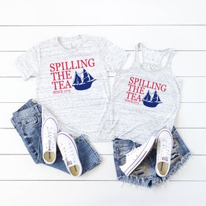 K&ouml;nnte beinhalten: Ein meliertes graues T-Shirt und Tanktop mit dem Text "Spilling the Tea Since 1773" in Rot und Blau, mit einer Segelboot-Grafik. Dazu blaue Jeans, Jeansshorts und wei&szlig;e Turnschuhe.