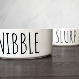 May include: Two white ceramic pet bowls with black lettering. The bowl on the left says "NIBBLE" and the bowl on the right says "SLURP".