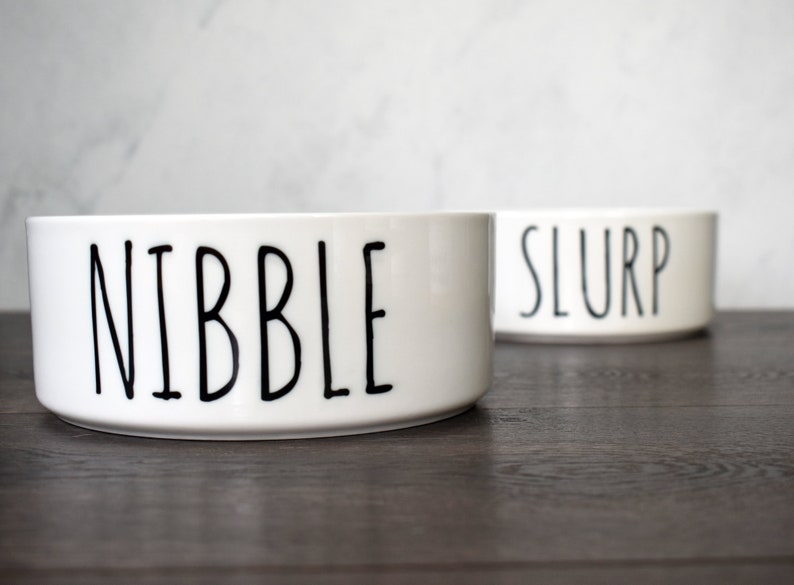 May include: Two white ceramic pet bowls with black lettering. The bowl on the left says "NIBBLE" and the bowl on the right says "SLURP".