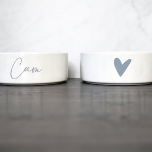 Puede incluir: Dos cuencos de cer&aacute;mica blanca para mascotas. Un cuenco tiene el nombre "Cam" escrito en &eacute;l en una fuente cursiva azul claro. El otro cuenco tiene un coraz&oacute;n azul claro.