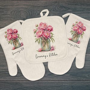 May include: A set of kitchen textiles featuring a watercolor floral design. The set includes two oven mitts and a square pot holder. Each item displays a bouquet of pink peonies in a glass jar, with the text "Granny's Kitchen".