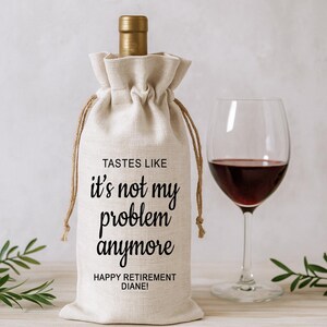 May include: A beige wine bottle bag with the text "TASTES LIKE it's not my problem anymore" and "HAPPY RETIREMENT DIANE!". A wine glass filled with red wine is next to the bag. The bag has a drawstring closure.