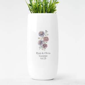 May include: A white ceramic vase with a floral design featuring purple and pink flowers and green leaves. The vase has the text "Ryan & Olivia Gusippe 9.9.19" printed on it. The vase is filled with green artificial grass.