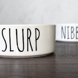 May include: Two white ceramic pet bowls with black lettering. The bowl on the left says "SLURP" and the bowl on the right says "NIBBLE".
