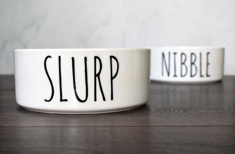 May include: Two white ceramic pet bowls with black lettering. The bowl on the left says "SLURP" and the bowl on the right says "NIBBLE".