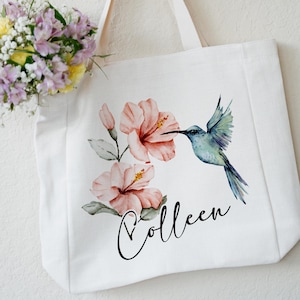 May include: White canvas tote bag with a watercolor design of pink hibiscus flowers and a blue hummingbird. The bag has the name "Colleen" written in black cursive.