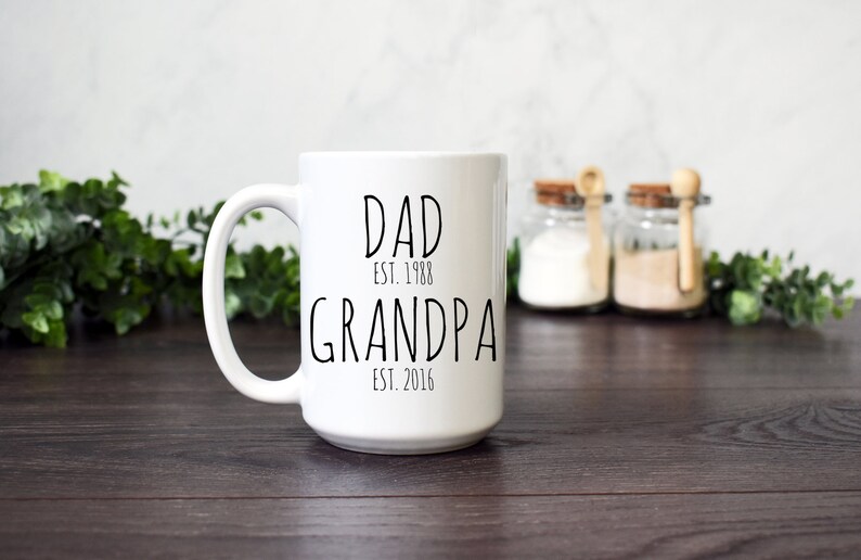 Może przedstawiać: Biały ceramiczny kubek z czarnym napisem "DAD EST. 1988 GRANDPA EST. 2016". Kubek ma duży uchwyt i stoi na ciemnej drewnianej powierzchni. W tle dekoracyjne słoiki i zieleń.