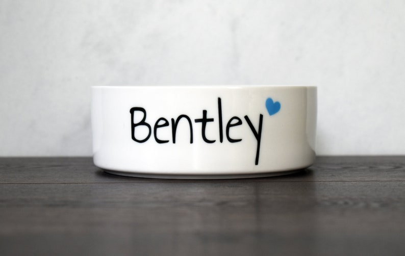 以下が含まれることがあります： 白いセラミック製のペットボウルに、黒い文字で「Bentley」と書いてあり、小さな青いハートが付いています。