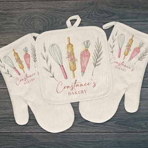 May include: A set of two beige oven mitts and a pot holder with a watercolor design of baking tools, including a rolling pin, whisk, and piping bag. The text "Constance's Bakery" is printed on each item.