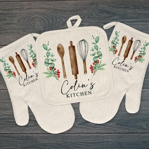 May include: Two off-white oven mitts and one square pot holder made of a linen-like fabric. Each piece features a watercolor illustration of a wooden spoon, rolling pin, whisk, and eucalyptus branches with red berries. The text Colin's KITCHEN is printed.