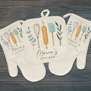 May include: Set of two kitchen oven mitts and a pot holder with a light brown background. The mitts and pot holder feature a watercolor design of kitchen utensils, including a rolling pin, whisk, spatula, and green leaves. The text "Maria's Kitchen" is printed on each item.