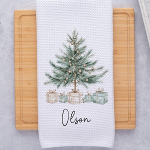May include: White waffle-weave kitchen towel with a watercolor Christmas tree design. The tree is decorated with gold ornaments and sits above wrapped gifts. The name "Olson" is written in script below the gifts.