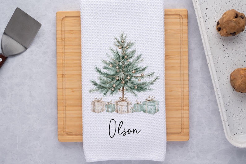 May include: White waffle-weave kitchen towel with a watercolor Christmas tree design. The tree is decorated with gold ornaments and sits above wrapped gifts. The name "Olson" is written in script below the gifts.