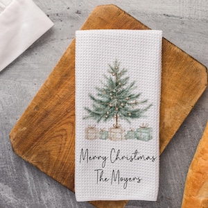 May include: White waffle-weave kitchen towel with a watercolor Christmas tree design and the text "Merry Christmas The Moyers." The towel features a green Christmas tree with ornaments and wrapped gifts. The towel is on a wooden cutting board.