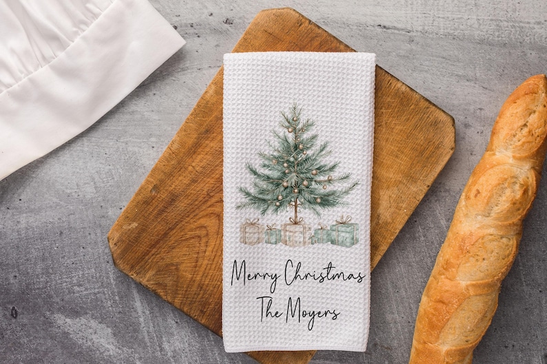 May include: White waffle-weave kitchen towel with a watercolor Christmas tree design and the text "Merry Christmas The Moyers." The towel features a green Christmas tree with ornaments and wrapped gifts. The towel is on a wooden cutting board.