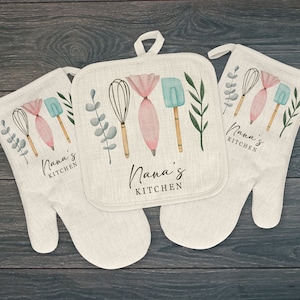 May include: Two off-white oven mitts and one square pot holder featuring a watercolor print of a whisk, a pink piping bag, a blue spatula, and green leaves. The text Nana's KITCHEN is printed in black on each item, which includes a hanging loop.