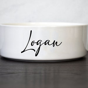 Puede incluir: Taz&oacute;n de cer&aacute;mica blanco para mascotas con el nombre "Logan" escrito en cursiva negra.