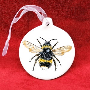 May include: White ceramic ornament with a hand-painted bumble bee with black and yellow stripes. The ornament has a white ribbon for hanging.
