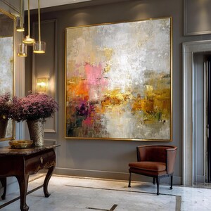 May include: Large abstract painting with a gold frame. The artwork features a blend of colors, including pink, orange, yellow, and gray, creating a textured effect. The painting hangs above a brown leather chair in a room with gold accents.