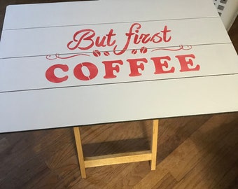 Puede incluir: Mesa plegable blanca con un letrero pintado de rojo que dice "But first coffee" con símbolos de granos de café.
