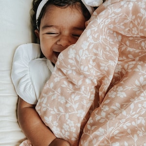 a happy baby wearing a white dress and headband bow giggling underneath her soft and lightweight fall swaddle muslin blanket in the magnolia tree peach color