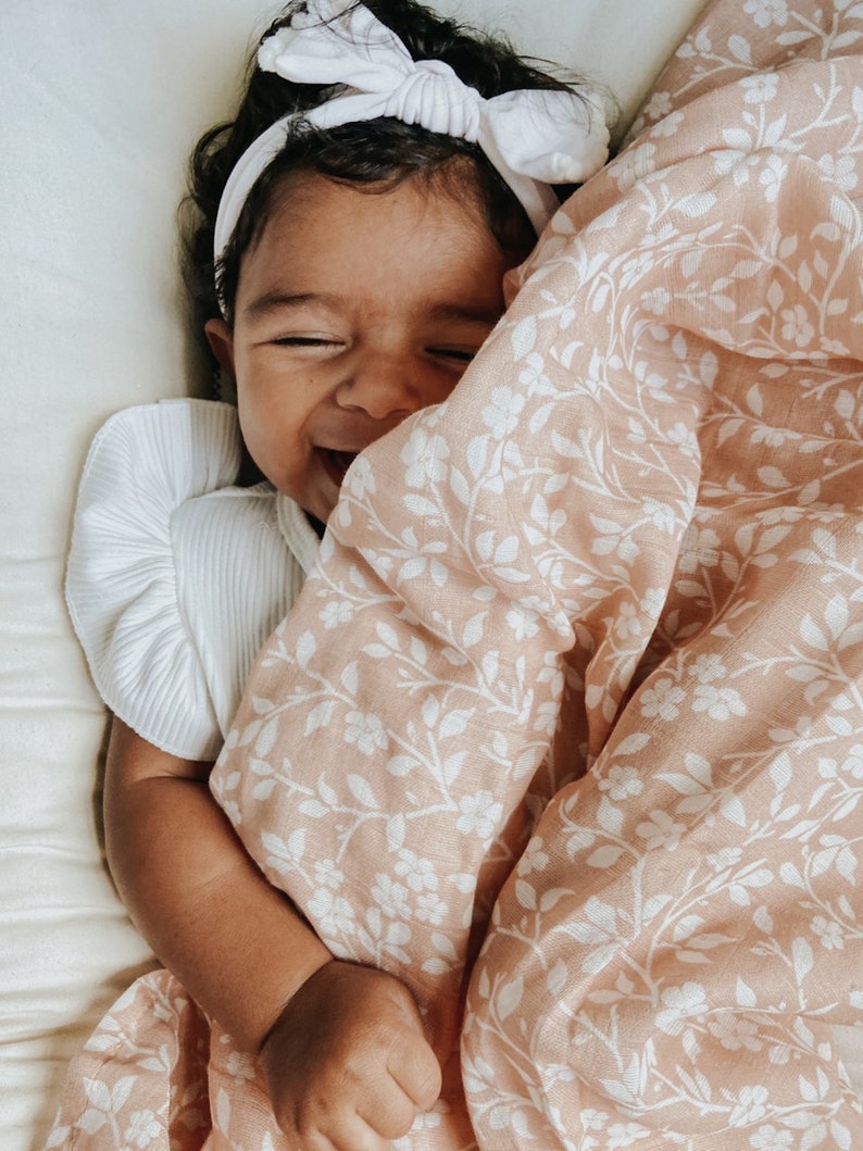 a happy baby wearing a white dress and headband bow giggling underneath her soft and lightweight fall swaddle muslin blanket in the magnolia tree peach color