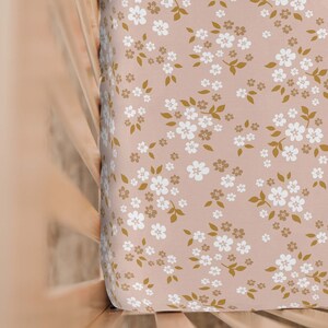 May include: A fitted crib sheet with a floral pattern. The sheet features a light pink background with a repeating pattern of white and gold-colored flowers and leaves. The sheet is made of soft fabric.