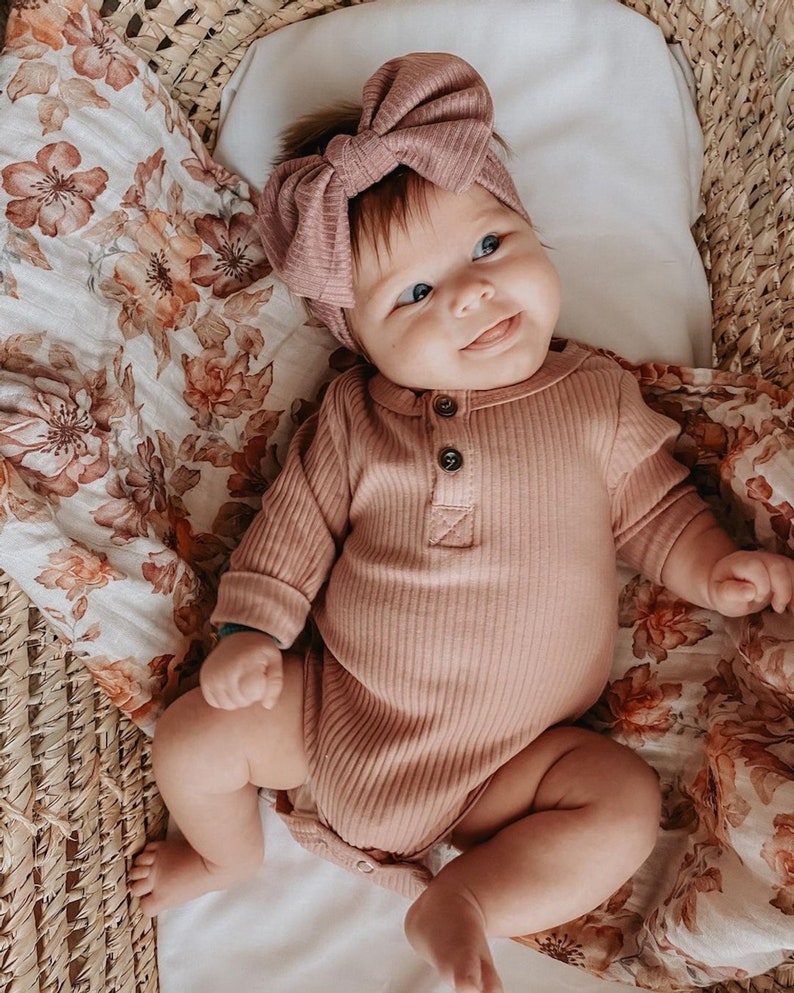an adorable little girl wearing neutral colored clothes and headband lying in her bassinet lined with the popular sunset floral white swaddle blanket for fall perfect for little girls