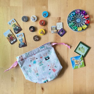 May include: A white drawstring bag with a bunny rabbit pattern. The bag is filled with small game pieces, including dice and cards. The cards feature illustrations of people and the text "Isle of Skye".