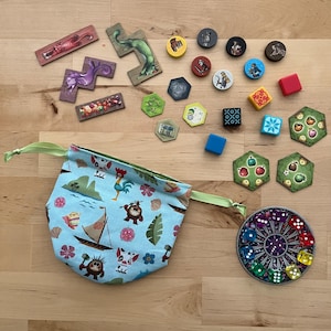 May include: A collection of board game components, including wooden tiles, tokens, dice, and a drawstring bag. The bag is light blue with a tropical print. Various game pieces are scattered on a wooden surface.