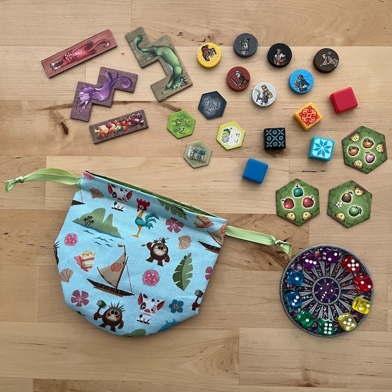 May include: A collection of board game components, including wooden tiles, tokens, dice, and a drawstring bag. The bag is light blue with a tropical print. Various game pieces are scattered on a wooden surface.