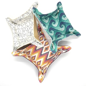 May include: Three square fabric trays with snap closures. One tray has a white background with black cartoon characters. Another has a teal wave pattern. The third has a colorful geometric design with orange, pink, and yellow.