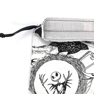 May include: A white drawstring bag featuring a black and white illustration of a skeleton character within a circular frame. The bag has a black drawstring and a white interior lining. The design is inspired by a well-known animated film.