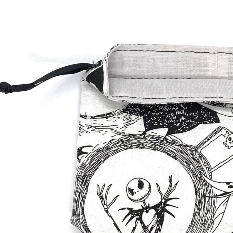 May include: A white drawstring bag featuring a black and white illustration of a skeleton character within a circular frame. The bag has a black drawstring and a white interior lining. The design is inspired by a well-known animated film.