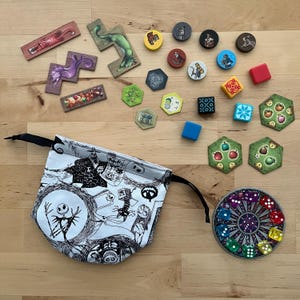 May include: A selection of board game pieces, including wooden tiles, circular tokens, dice, and a drawstring bag. The bag has a black and white illustration. The game pieces are in various colours.
