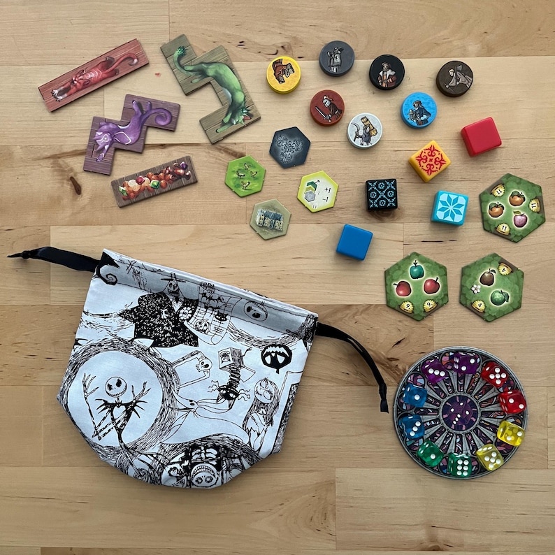 May include: A selection of board game pieces, including wooden tiles, circular tokens, dice, and a drawstring bag. The bag has a black and white illustration. The game pieces are in various colours.