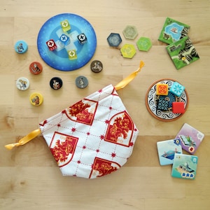 May include: A collection of board game components, including a drawstring bag with a red and gold crest pattern, game tiles, dice, and player tokens. The bag is tied with a yellow ribbon.